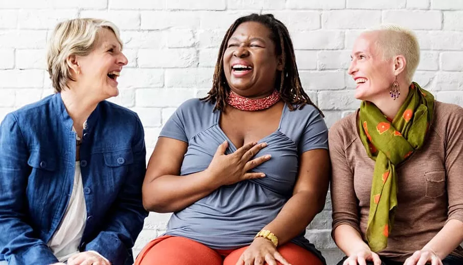 Three women of different ethnicities sit together against a white brick wall, laughing joyfully in casual, colorful clothing.