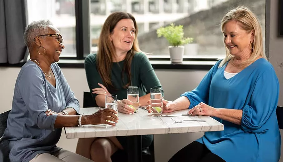 Three women of varying ages sit around a small marble table in a bright restaurant, smiling and conversing over glasses of water with lime.