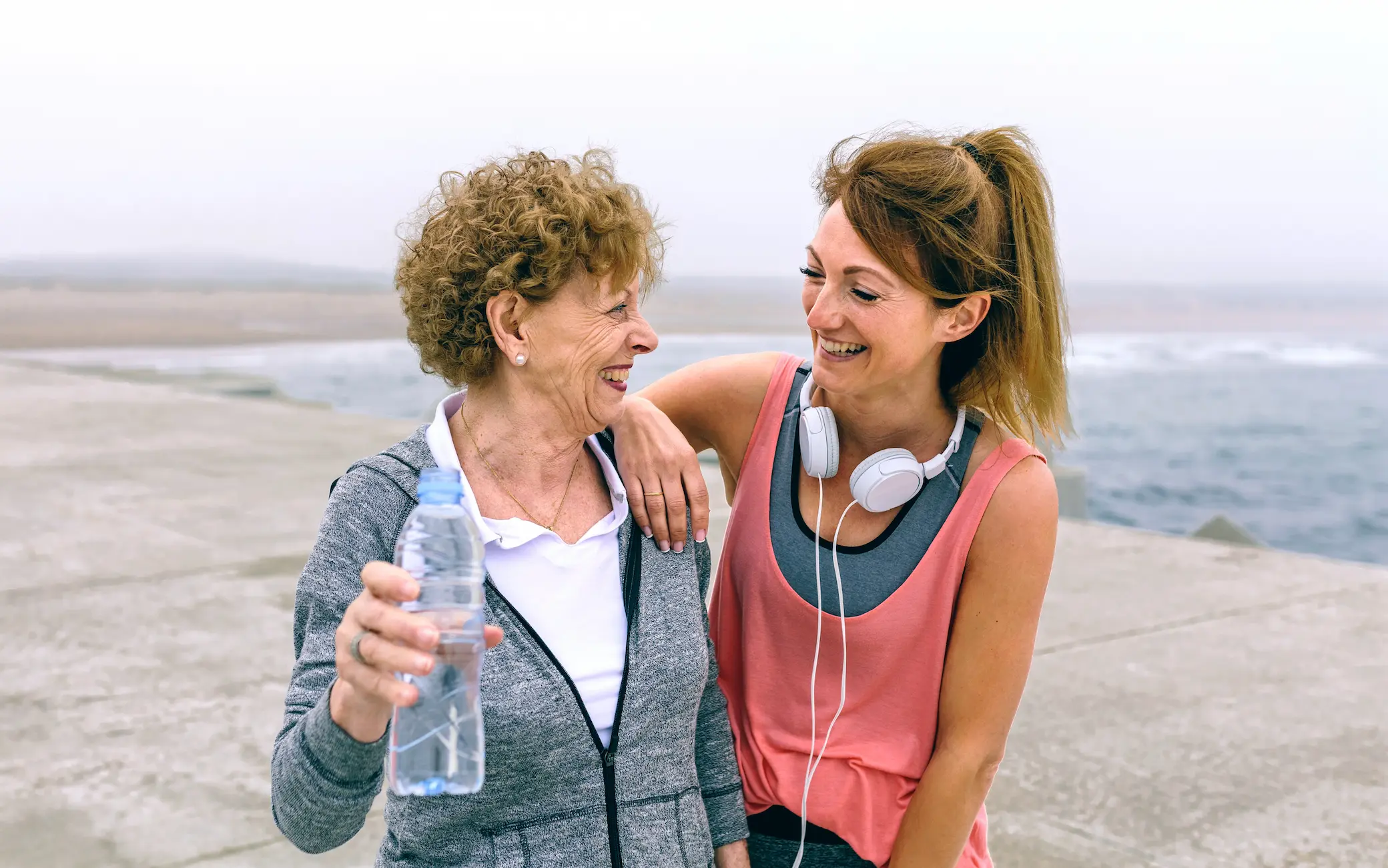 Two women in athletic wear smile at each other near a beach. An older woman holds a water bottle; a younger woman has headphones around her neck.