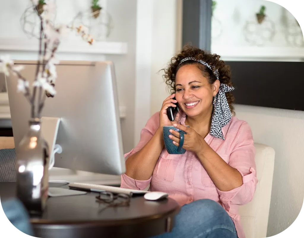 Smiling woman talking on the phone holding a mug in front of a desktop computer