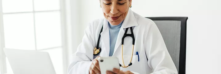 A female doctor in a white lab coat and stethoscope smiles while looking at her smartphone, seated at a desk in a bright clinical office.