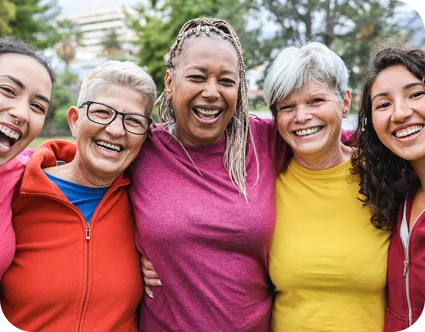 Five diverse women of varying ages are smiling and laughing together, arms around each other, wearing colorful athletic tops outdoors.
