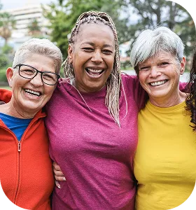 Three older women smiling and laughing together outdoors, arms around each other, wearing colorful tops (red, pink, yellow), with greenery in the background.