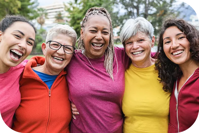 Five diverse women of varying ages are smiling and laughing together, arms around each other, wearing colorful athletic wear outdoors with trees in the background.