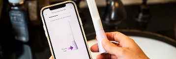 A person holds a pregnancy test stick next to a smartphone displaying an app with a purple triangle graphic, near a bathroom sink.