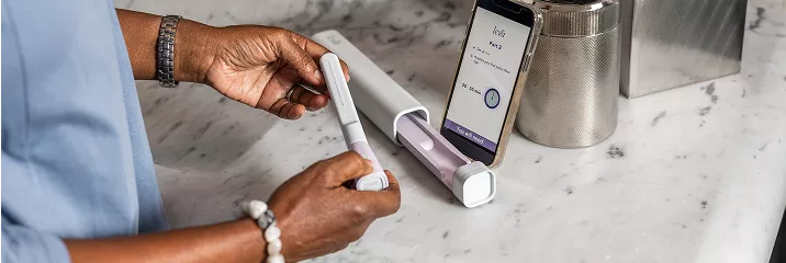 A person's hands are inserting a test strip into a medical device on a marble countertop, with a smartphone displaying the **Invia** app beside it.