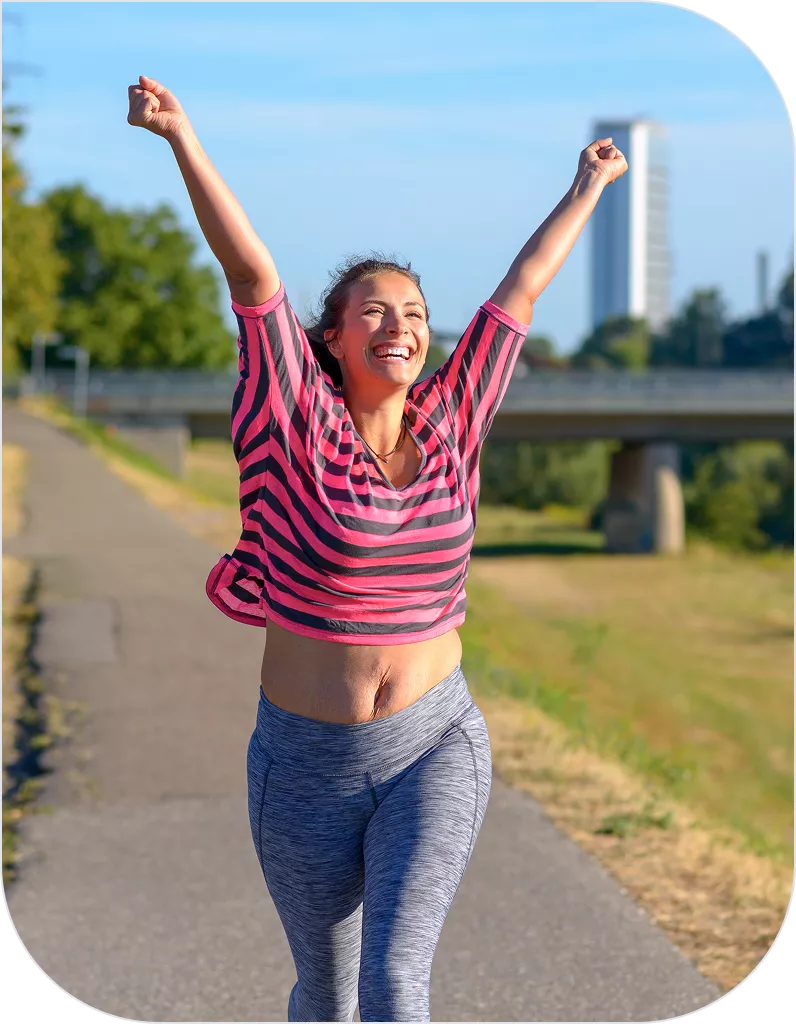 A young woman in a pink striped top and gray leggings raises both arms triumphantly while smiling broadly on an outdoor path near a bridge.