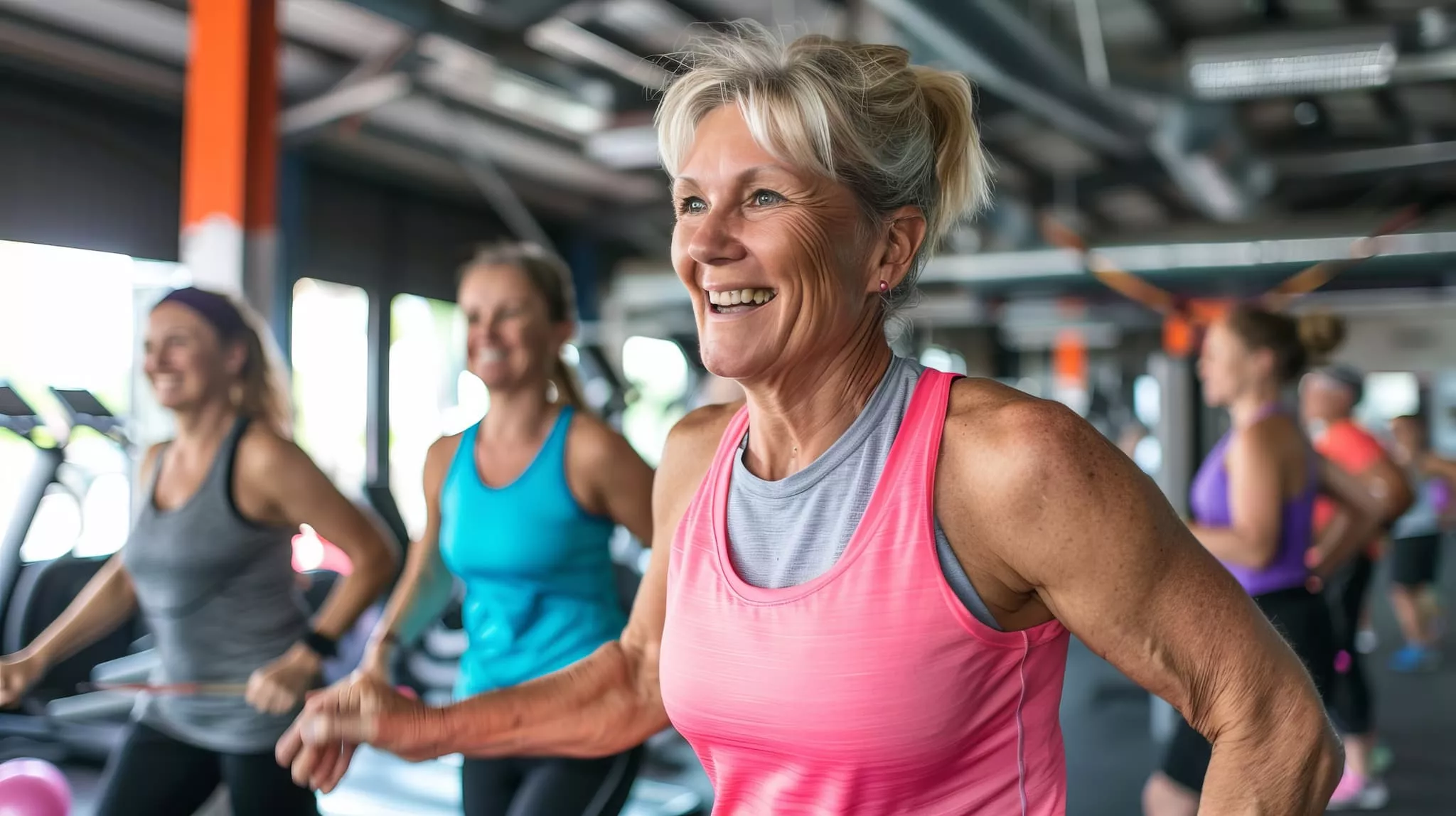 A middle-aged woman in a pink tank top smiles brightly while exercising in a busy gym, with several other women working out in the background.