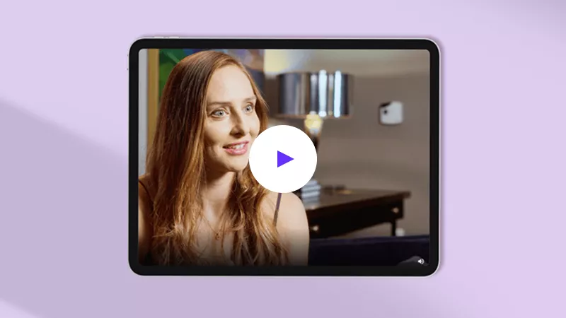 A woman with long auburn hair smiles warmly, displayed on a tablet screen with a play button overlay, set against a lavender background.