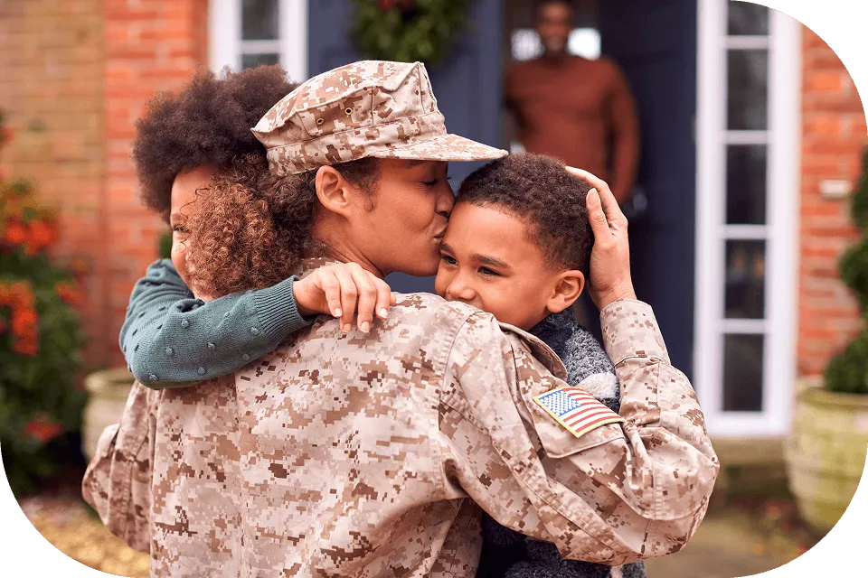 A female soldier in US military camouflage uniform embraces and kisses two young children outside a brick home, with a person visible in the doorway behind them.