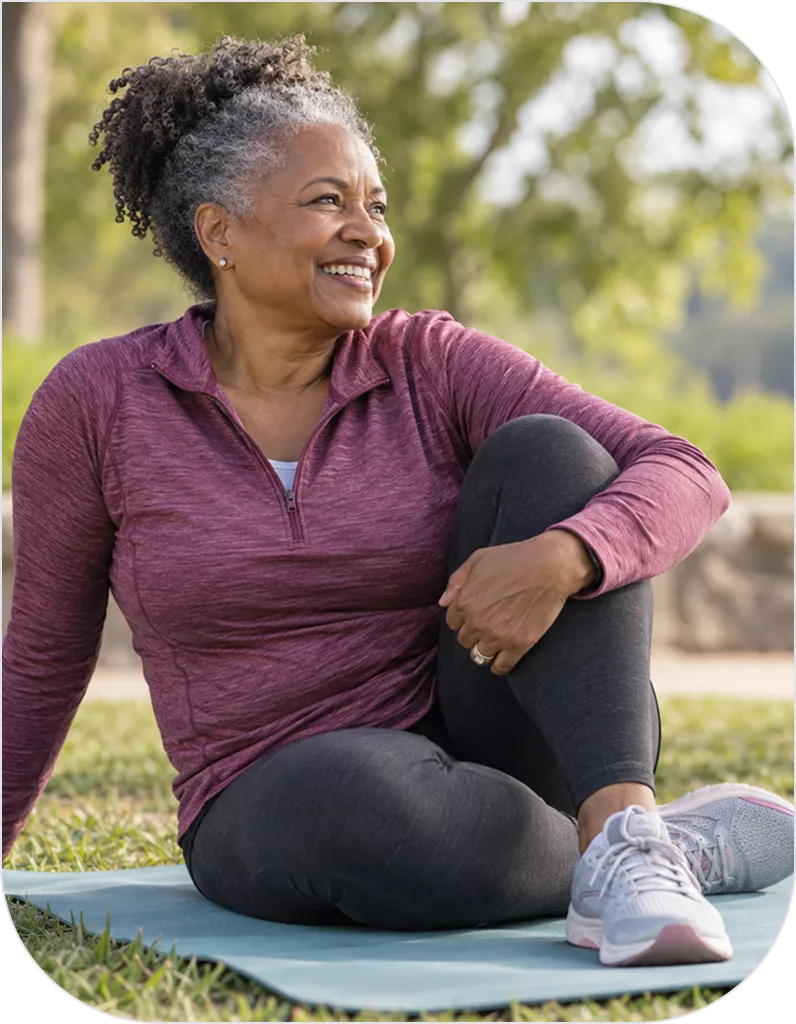 A middle-aged woman sits relaxed on a yoga mat outdoors, smiling, wearing a maroon athletic top, black leggings, and gray sneakers, with green trees behind her.